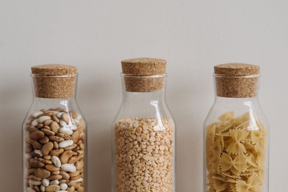 Three clear glass jars with cork lids hold beans, lentils, and pasta showcasing pantry organization.
