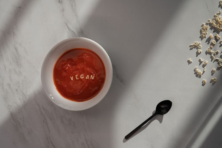 A bowl of vegan tomato soup with pasta letters on a marble surface in natural sunlight.