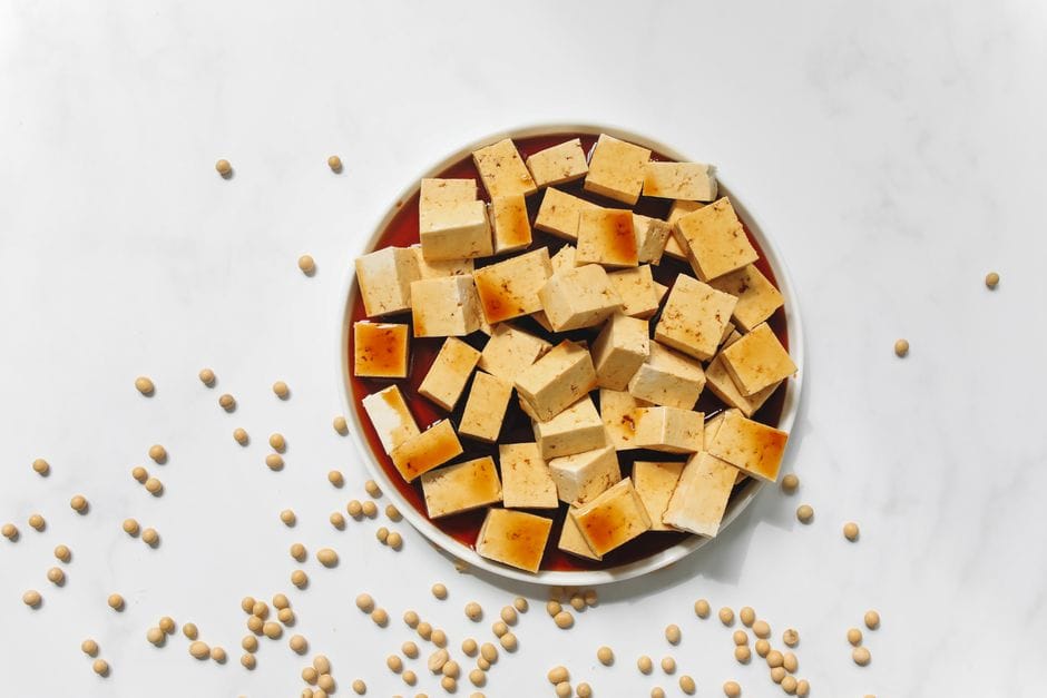 Top view of cubed tofu on a plate with scattered soybeans on a white marble surface.