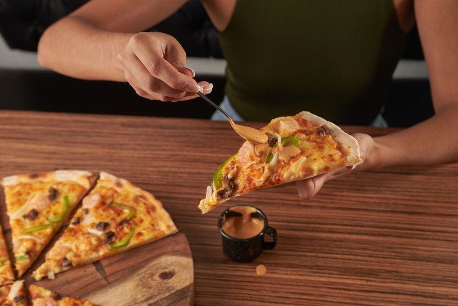 Person adding sauce to a slice of pizza on a wooden table. Perfect for food lovers.