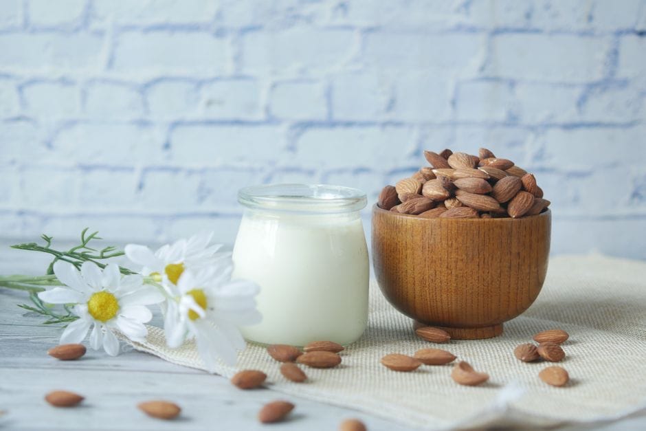 Natural almonds in a wooden bowl with a glass jar of milk and white chamomile flowers on a wood table.