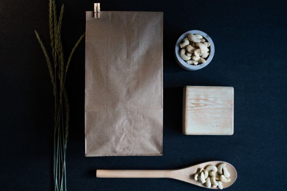 Minimalist flat lay featuring a brown paper bag, cashews, spoon, and wooden block on dark background.