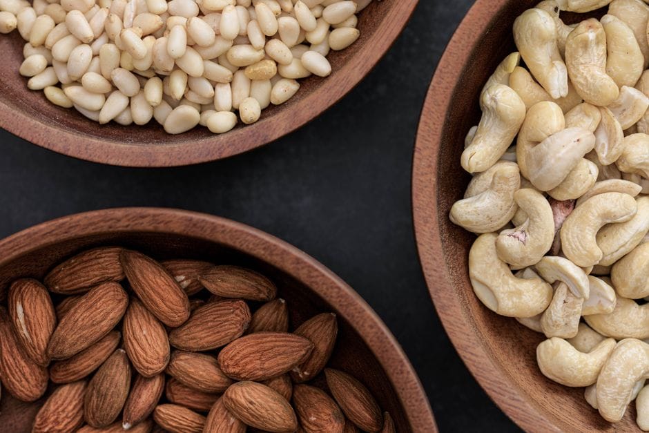 Top view of almonds, cashews, and pine nuts in wooden bowls against a dark backdrop.