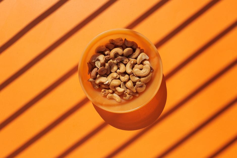 Flat lay image of cashew nuts in an orange bowl on a striped orange background, perfect for organic food themes.