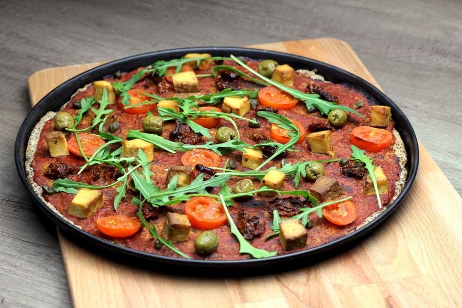 Vegan pizza topped with cherry tomatoes, arugula, tofu, olives, and vegan cheese on a wooden board.