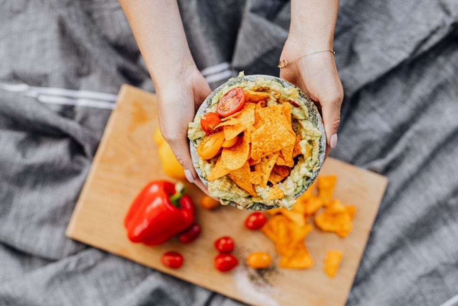 Close-up of hands holding nachos with guacamole and cherry tomatoes over a cutting board.