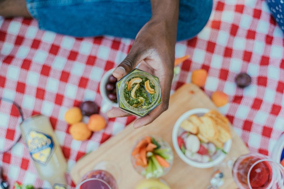 Close-up of a hand holding a jar of cashew pesto on a picnic blanket, highlighting a summer outdoor vibe.