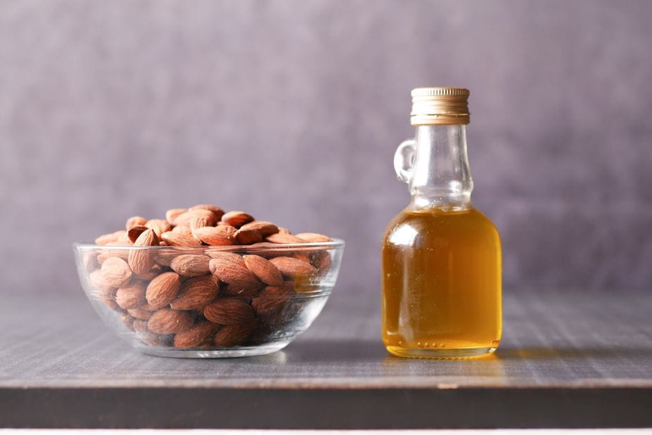 A close-up of almonds in a glass bowl alongside a bottle of almond oil.